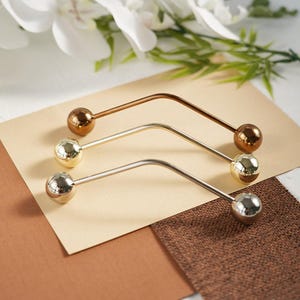 Pu&ograve; includere: Tre spille decorative per hijab in oro, argento e bronzo. Ogni spilla presenta una barra curva con una sfera sfaccettata a ciascuna estremit&agrave;. Le spille sono esposte su uno sfondo neutro con accenti floreali, evidenziando il loro design elegante.