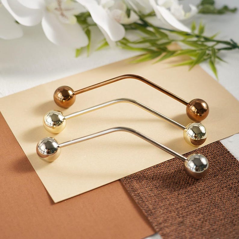 Pu&ograve; includere: Tre spille decorative per hijab in oro, argento e bronzo. Ogni spilla presenta una barra curva con una sfera sfaccettata a ciascuna estremit&agrave;. Le spille sono esposte su uno sfondo neutro con accenti floreali, evidenziando il loro design elegante.