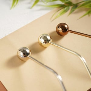 Pu&ograve; includere: Tre forcine per capelli in metallo con la parte superiore sferica. Una &egrave; argentata, una dorata e una color bronzo. Le forcine sono disposte diagonalmente su una superficie marrone chiaro, con fogliame verde sullo sfondo.