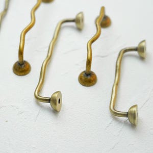 K&ouml;nnte beinhalten: Nahaufnahme mehrerer antiker Messing-Schrankgriffe. Die Griffe haben ein wellenf&ouml;rmiges, gebogenes Design mit rundem Sockel und einem flachen, kreisf&ouml;rmigen Ende. Das Messing hat eine warme, gealterte Patina.