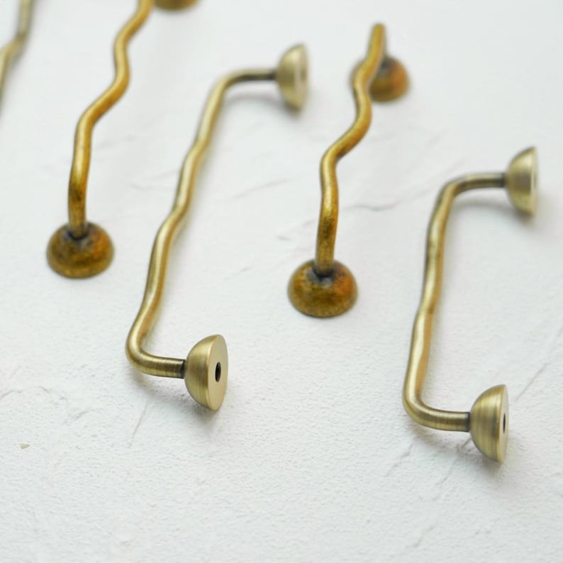 K&ouml;nnte beinhalten: Nahaufnahme mehrerer antiker Messing-Schrankgriffe. Die Griffe haben ein wellenf&ouml;rmiges, gebogenes Design mit rundem Sockel und einem flachen, kreisf&ouml;rmigen Ende. Das Messing hat eine warme, gealterte Patina.