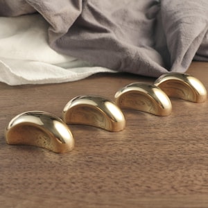 Puede incluir: Cuatro objetos dorados en forma de media luna están dispuestos en fila sobre una superficie de madera. Los objetos tienen una superficie lisa y reflectante, y están colocados frente a una tela gris y blanca.