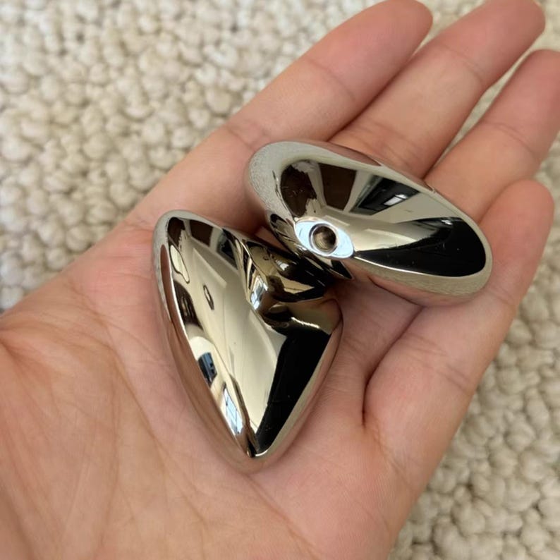 May include: Two polished, heart-shaped, silver-colored metal objects. The objects are smooth and reflective, with a small hole in one. The objects are held in a hand.