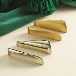 May include: Four contemporary cabinet pulls in gold and silver finishes. The pulls feature a curved, ergonomic shape. A green fabric and tassel are in the background.