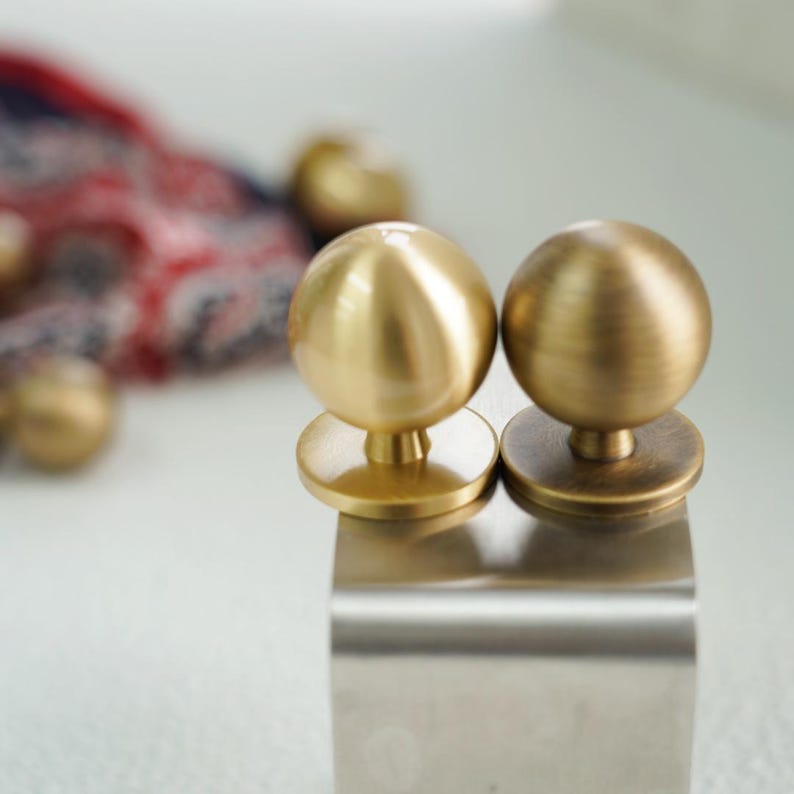 Op de afbeelding: Twee goudkleurige, bolvormige kastknoppen met een platte basis. De knoppen staan op een geborsteld metalen oppervlak. De achtergrond is wazig, met hints van een patroonstof in rood, wit en blauw.