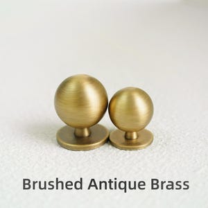 Op de afbeelding: Twee meubelknoppen van geborsteld antiek messing. Elke knop heeft een bolvormige vorm op een cirkelvormige basis. De tekst "Brushed Antique Brass" staat eronder.