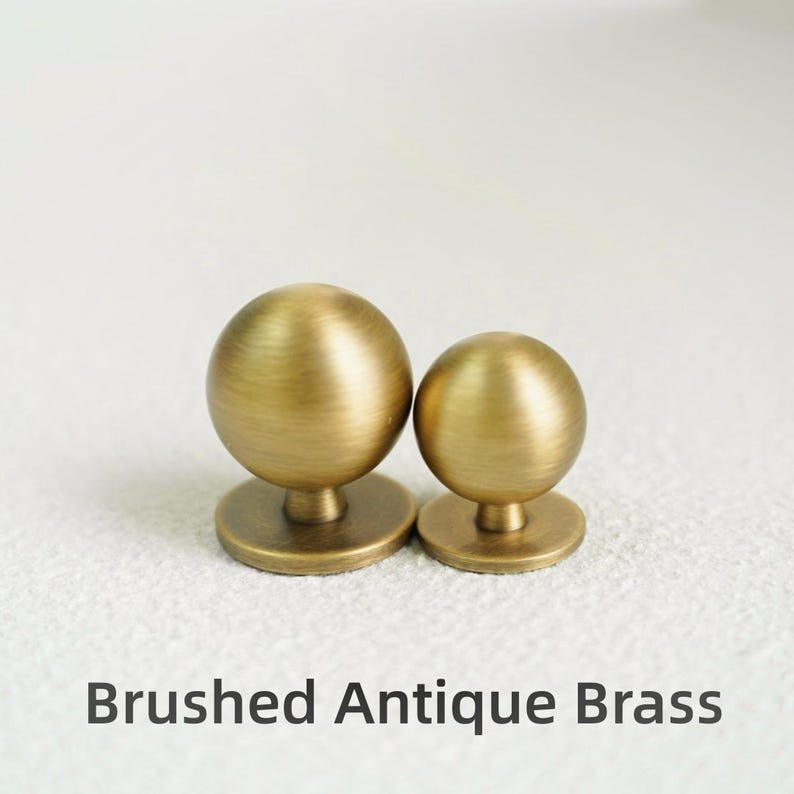 Op de afbeelding: Twee meubelknoppen van geborsteld antiek messing. Elke knop heeft een bolvormige vorm op een cirkelvormige basis. De tekst "Brushed Antique Brass" staat eronder.