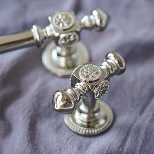 以下が含まれることがあります： 装飾的なディテールが施された、磨かれたシルバーのキャビネットノブが2つ。各ノブは、中央に花の模様と装飾的なベースが特徴です。ノブは、柔らかいラベンダー色の生地の上に置かれ、その反射面を際立たせています。