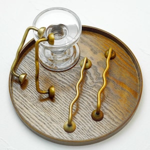 K&ouml;nnte beinhalten: Ein klarer Glaskerzenhalter mit rundem Sockel und Messinggriff sowie zwei wellenf&ouml;rmige Messinggriffe sind auf einem runden Holztablett angeordnet. Die Griffe haben ein einzigartiges, k&uuml;nstlerisches Design und verleihen einen Hauch von Eleganz.