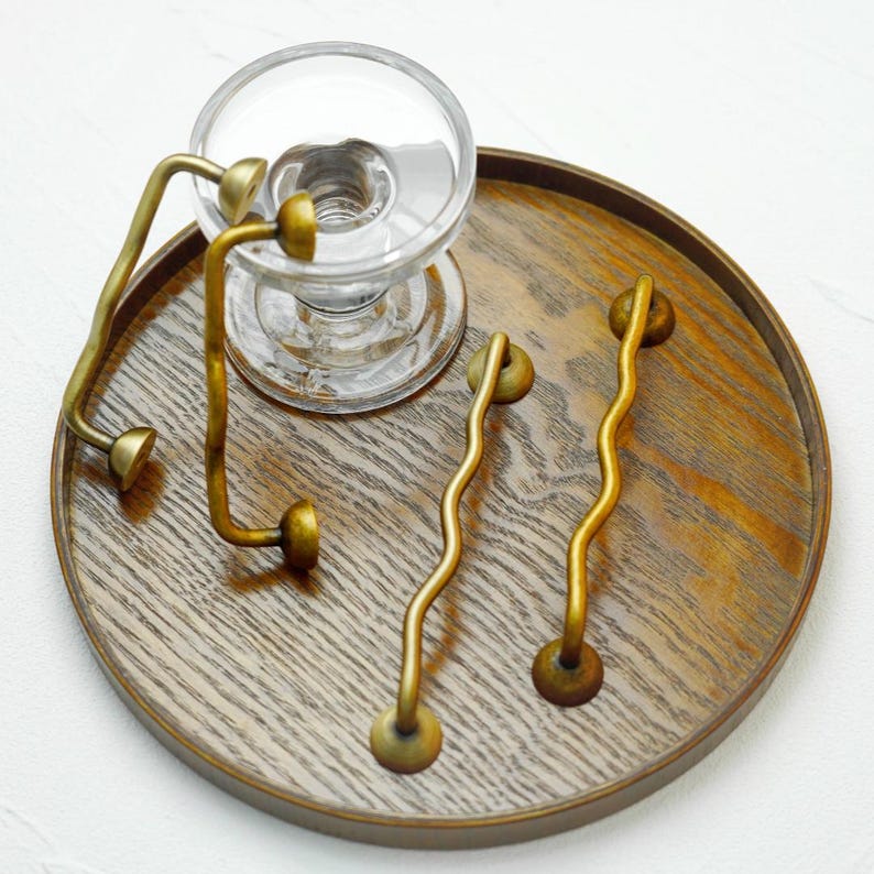 K&ouml;nnte beinhalten: Ein klarer Glaskerzenhalter mit rundem Sockel und Messinggriff sowie zwei wellenf&ouml;rmige Messinggriffe sind auf einem runden Holztablett angeordnet. Die Griffe haben ein einzigartiges, k&uuml;nstlerisches Design und verleihen einen Hauch von Eleganz.
