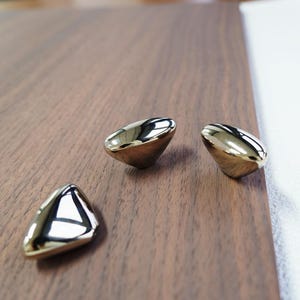 May include: Three shiny, gold-colored cabinet knobs with a triangular shape are arranged on a dark brown wooden surface. The knobs reflect light, creating a bright, polished appearance. The hardware is likely for furniture or home decor.