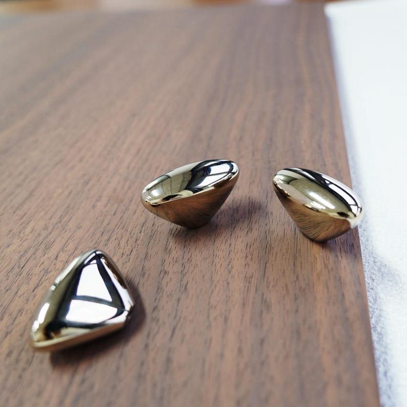 May include: Three shiny, gold-colored cabinet knobs with a triangular shape are arranged on a dark brown wooden surface. The knobs reflect light, creating a bright, polished appearance. The hardware is likely for furniture or home decor.