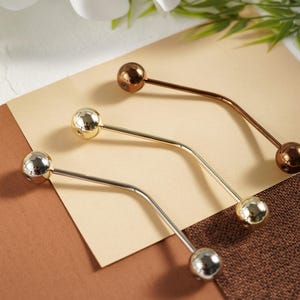 Pu&ograve; includere: Tre spille per scialle in metallo argentato, dorato e bronzo. Ogni spilla presenta una barra curva con una sfera decorativa a ciascuna estremit&agrave;. Le spille sono disposte su uno sfondo neutro con una combinazione di colori marrone e beige.