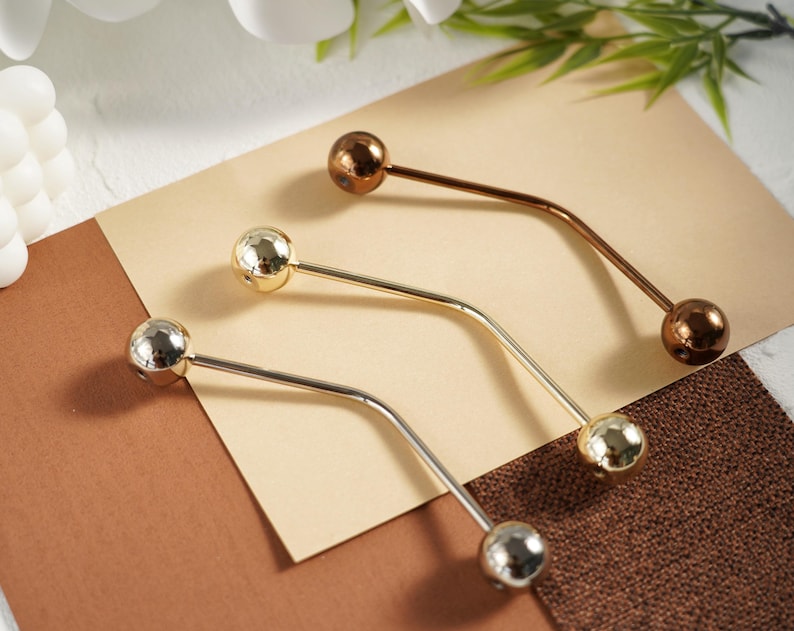 Pu&ograve; includere: Tre spille per scialle in metallo argentato, dorato e bronzo. Ogni spilla presenta una barra curva con una sfera decorativa a ciascuna estremit&agrave;. Le spille sono disposte su uno sfondo neutro con una combinazione di colori marrone e beige.