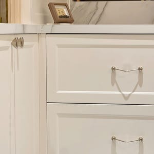 Pu&ograve; includere: Armadietti da cucina bianchi con ferramenta argentata. Gli armadi hanno un design pulito e moderno con una finitura liscia e verniciata. Una piccola immagine incorniciata &egrave; appoggiata sul piano di lavoro. La ferramenta comprende pomelli e maniglie.
