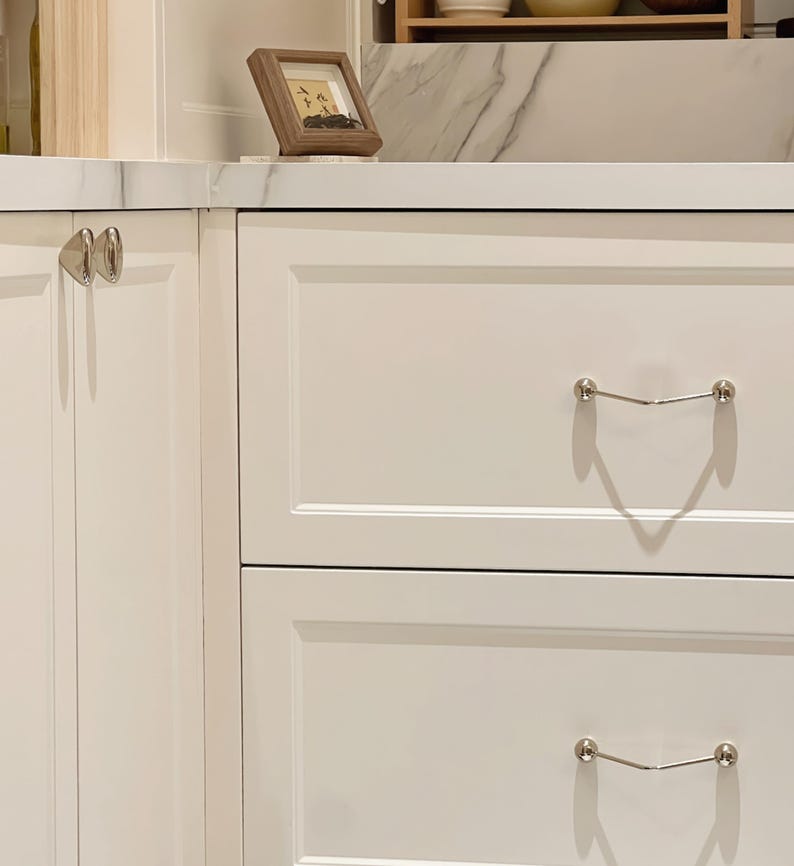 Pu&ograve; includere: Armadietti da cucina bianchi con ferramenta argentata. Gli armadi hanno un design pulito e moderno con una finitura liscia e verniciata. Una piccola immagine incorniciata &egrave; appoggiata sul piano di lavoro. La ferramenta comprende pomelli e maniglie.