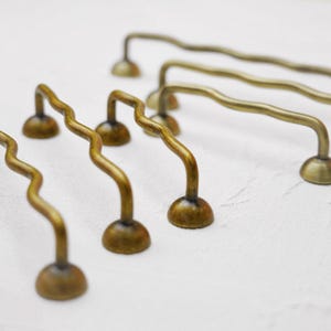 K&ouml;nnte beinhalten: Eine Sammlung antiker Messing-Schrankgriffe mit einem einzigartigen, wellenf&ouml;rmigen Design. Die Griffe haben einen gebogenen Griff und sind auf einer abgerundeten Basis montiert. Die Messingoberfl&auml;che verleiht ihnen einen Vintage-Look, ideal f&uuml;r M&ouml;belrestaurierung.