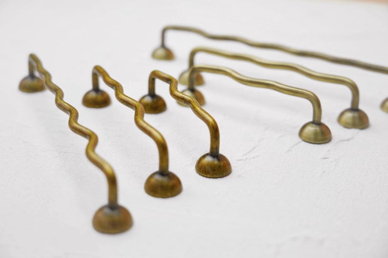 K&ouml;nnte beinhalten: Eine Sammlung antiker Messing-Schrankgriffe mit einem einzigartigen, wellenf&ouml;rmigen Design. Die Griffe haben einen gebogenen Griff und sind auf einer abgerundeten Basis montiert. Die Messingoberfl&auml;che verleiht ihnen einen Vintage-Look, ideal f&uuml;r M&ouml;belrestaurierung.