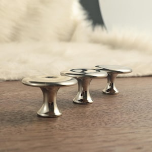 Op de afbeelding: Drie glanzende, zilverkleurige kastknoppen met een paddenstoelvormig ontwerp zijn gerangschikt op een houten oppervlak. De knoppen hebben een gladde, reflecterende afwerking en een klassieke uitstraling. De achtergrond heeft een zacht, wit, getextureerd oppervlak.