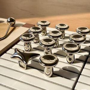 May include: A collection of shiny, silver mushroom-shaped cabinet knobs and a star-shaped knob. The knobs are arranged on a white and gray tiled surface. The mushroom knobs have a rounded top and a cylindrical base.
