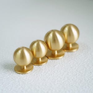 Op de afbeelding: Vier messing kastknoppen met een bolvormig ontwerp. De knoppen zijn diagonaal gerangschikt, elk met een cirkelvormige basis en een gladde, gepolijste afwerking. De knoppen hebben een warme goudkleur.