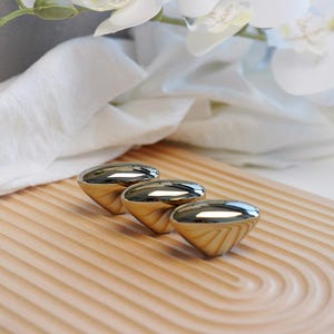 May include: Three gold-colored cabinet knobs with a reflective, polished finish are arranged on a light brown, wooden tray with a grooved design. White flowers and a white cloth are in the background.