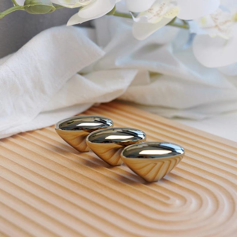 May include: Three gold-colored cabinet knobs with a reflective, polished finish are arranged on a light brown, wooden tray with a grooved design. White flowers and a white cloth are in the background.