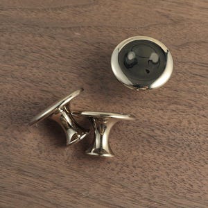 May include: Three gold-colored cabinet knobs with a mushroom-shaped design. One knob is round with a flat top, while the other two have a flared base and a flat, circular top. The knobs are displayed on a wooden surface.