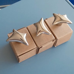 Puede incluir: Tres tiradores de armario plateados pulidos en forma de estrella, presentados sobre pequeñas cajas de cartón marrón. Los tiradores tienen una superficie lisa y reflectante, sobre un fondo azul claro. El diseño de estrella añade un toque moderno.
