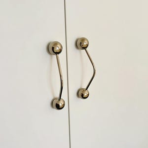 Pu&ograve; includere: Due maniglie per ante di armadi. Ogni maniglia ha una finitura cromata lucida, con un pomello sferico a ciascuna estremit&agrave; collegato da un gambo curvo e sottile. Le maniglie sono montate su un'anta bianca dell'armadio.