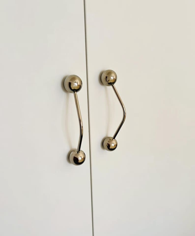 Pu&ograve; includere: Due maniglie per ante di armadi. Ogni maniglia ha una finitura cromata lucida, con un pomello sferico a ciascuna estremit&agrave; collegato da un gambo curvo e sottile. Le maniglie sono montate su un'anta bianca dell'armadio.