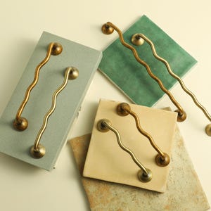 Può includere: Quattro pomelli decorativi ondulati in ottone sono esposti su varie piastrelle colorate. I pomelli hanno una forma organica unica e sono fissati con hardware rotondo e decorativo. I pomelli sono di un caldo colore oro.