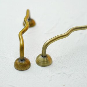 K&ouml;nnte beinhalten: Zwei antike Messing-Schrankgriffe mit wellenf&ouml;rmigem, geschwungenem Design. Jeder Griff hat einen abgerundeten Sockel und einen geschwungenen Griff, was einen einzigartigen und eleganten Look erzeugt. Das Messing hat eine warme, gealterte Patina.
