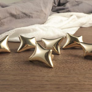 Puede incluir: Seis tiradores dorados en forma de estrella colocados sobre una superficie de madera. Los tiradores tienen un acabado liso y reflectante y un diseño geométrico único. Un tejido suave en tonos grises y blancos se encuentra en el fondo.