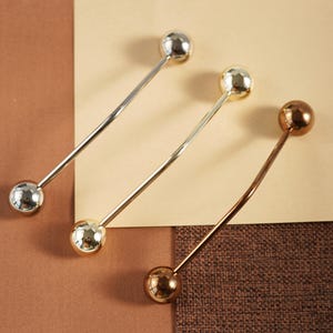 Pu&ograve; includere: Tre barrette per colletto in metallo argentato, dorato e bronzo. Ogni barretta ha una sfera sferica su ciascuna estremit&agrave; e una barretta dritta o curva che le collega. Le barrette sono disposte su una superficie marrone e beige.