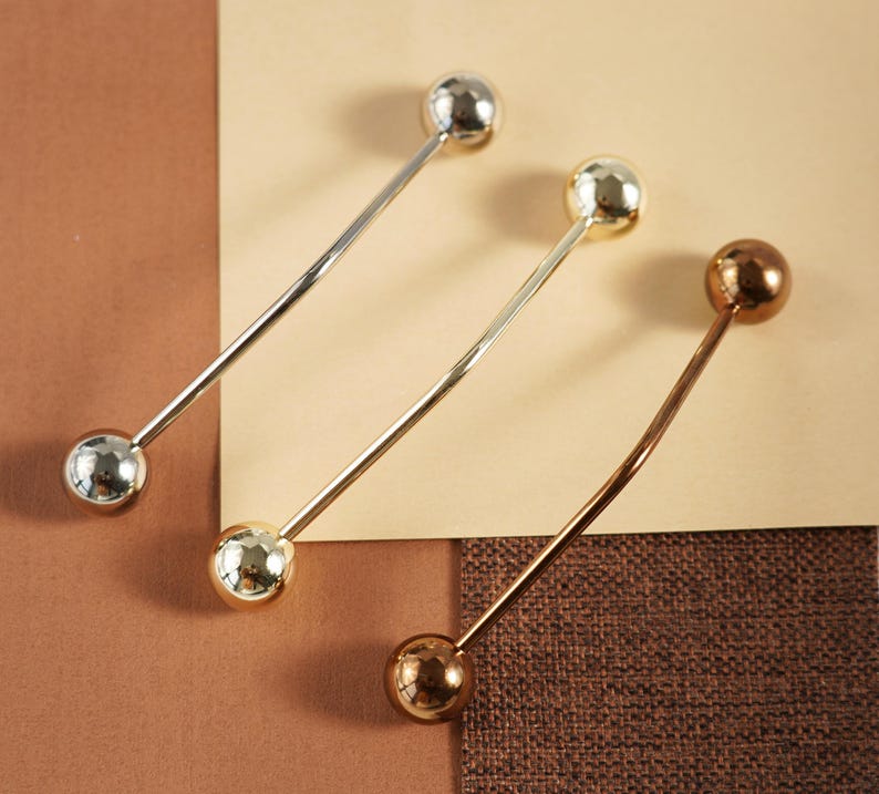 Pu&ograve; includere: Tre barrette per colletto in metallo argentato, dorato e bronzo. Ogni barretta ha una sfera sferica su ciascuna estremit&agrave; e una barretta dritta o curva che le collega. Le barrette sono disposte su una superficie marrone e beige.