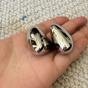 May include: Two polished, teardrop-shaped, silver-colored objects. The objects are smooth and reflective, with a small opening at the top. They are held in an open hand.