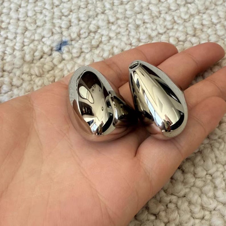 May include: Two polished, teardrop-shaped, silver-colored objects. The objects are smooth and reflective, with a small opening at the top. They are held in an open hand.