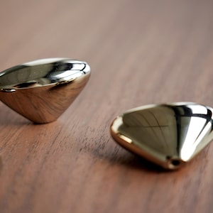 May include: Three gold-colored, teardrop-shaped cabinet knobs with a polished, reflective surface. The knobs are arranged on a dark brown wooden surface, creating a contrast in textures and colors. Hardware for home decor.