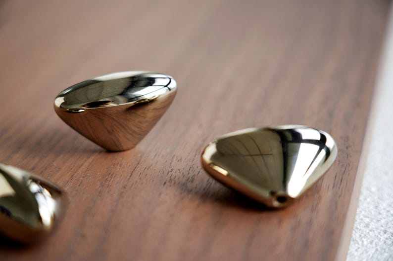 May include: Three gold-colored, teardrop-shaped cabinet knobs with a polished, reflective surface. The knobs are arranged on a dark brown wooden surface, creating a contrast in textures and colors. Hardware for home decor.
