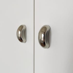 Könnte beinhalten: Zwei polierte Chrom-Schrankgriffe an einer weißen Schranktür. Die Griffe haben ein gebogenes, ergonomisches Design, das die Umgebung widerspiegelt. Die Hardware ist ein moderner Akzent für Küchen- oder Badezimmerschränke.