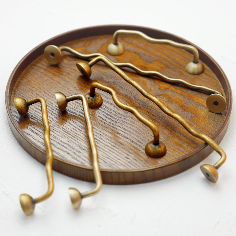 K&ouml;nnte beinhalten: Eine Sammlung von messingfarbenen Schrankgriffen und -kn&ouml;pfen mit wellenf&ouml;rmigem Design. Die Beschl&auml;ge sind auf einem runden, dunklen Holztablett angeordnet, was ihren einzigartigen, dekorativen Stil zeigt. Die Griffe haben eine Vintage-&Auml;sthetik.