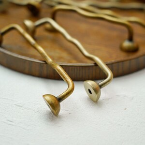 K&ouml;nnte beinhalten: Nahaufnahme mehrerer antiker Messing-Schrankgriffe. Die Griffe haben ein gebogenes Design mit einem abgerundeten Ende und einer Montagebasis. Das Messing hat eine warme, gealterte Patina. Die Griffe sind auf einem Holztablett angeordnet.