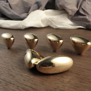 May include: Six gold-colored, teardrop-shaped cabinet knobs. The knobs are arranged on a wooden surface, reflecting light. The knobs have a smooth, polished finish, and are likely made of metal.