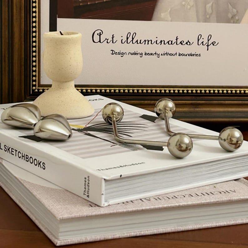 Pu&ograve; includere: Una natura morta con un portacandele beige, oggetti decorativi argentati e una pila di libri. L'opera d'arte incorniciata sopra recita "Art illuminates life". Gli oggetti sono disposti su una superficie bianca e beige.