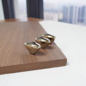 May include: Three gold-colored cabinet knobs with a rounded, teardrop shape are arranged on a dark brown wooden surface. The knobs have a shiny, reflective finish. The background is blurred, showing a window and a cityscape.