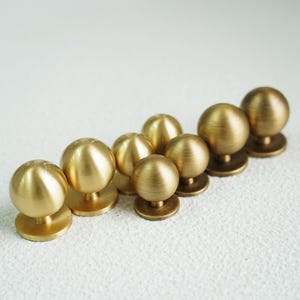 Op de afbeelding: Een verzameling van acht messing kastknoppen. Elke knop heeft een ronde, geborstelde messing bol bovenop een cirkelvormige basis. De knoppen zijn in een diagonale lijn gerangschikt, wat hun eenvoudige, maar elegante ontwerp laat zien. De warme goudkleur voegt een vleugje verfijning toe.