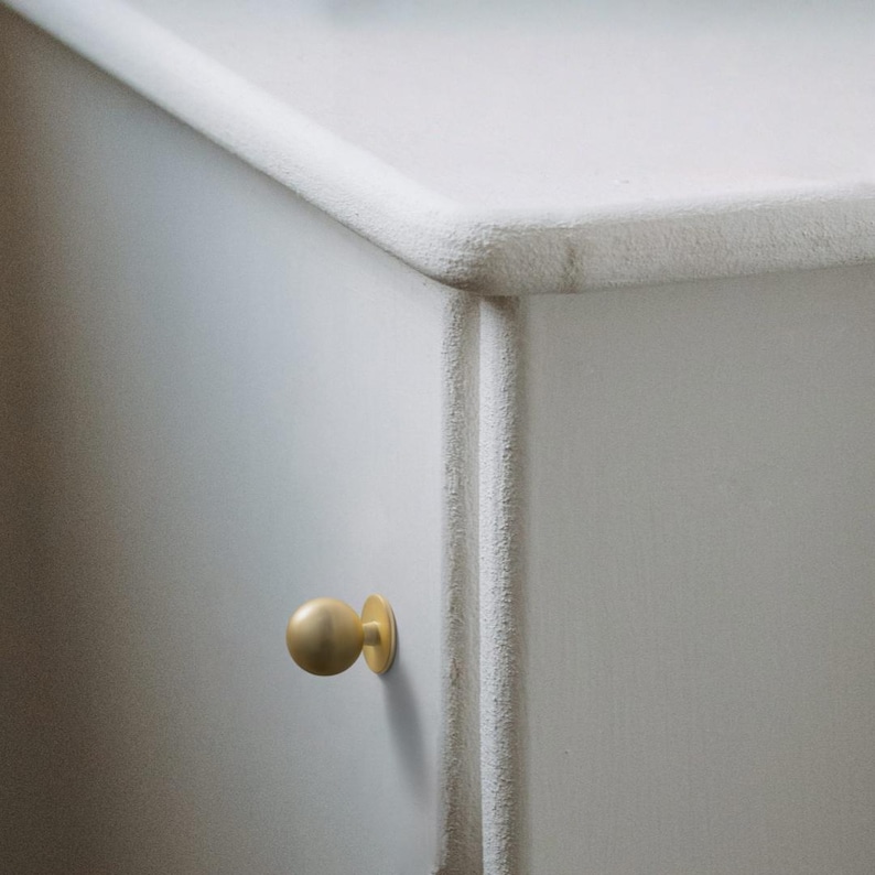 Op de afbeelding: Close-up van een deel van een wit geschilderde kast met een afgeronde rand en een goudkleurige bolvormige knop. De kast heeft een getextureerd oppervlak en een verticale decoratieve afwerking.