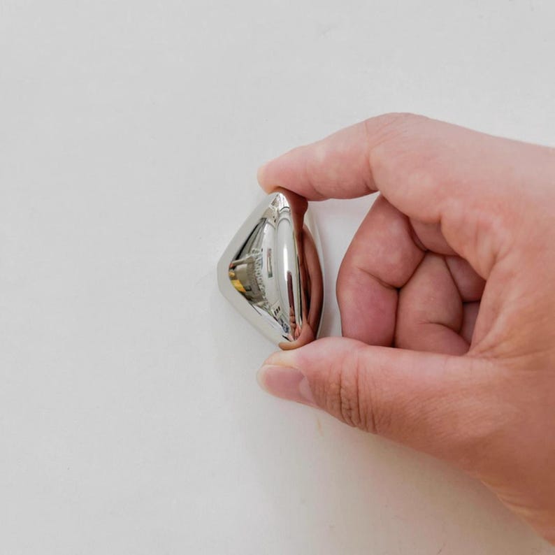 May include: A small, shiny, silver-colored object with a rounded, triangular shape. The object has a reflective surface and appears to be made of metal. The item is held between fingers against a white background.