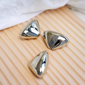 May include: Three shiny, metallic, triangular objects with rounded edges are arranged on a light-colored wooden surface. The objects reflect the surrounding environment, creating a mirrored effect. A white cloth is in the background.